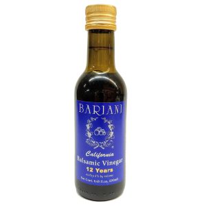 Label of Bariani Aged Balsamic Vinegar bottle showing product description. It describes the vinegar as traditionally made from cooked grape must, slowly fermented and aged in wooden barrels. The vinegar has a dark brown color, intense woody flavor, and sweet mature aroma. Ingredients listed: cooked grape must. It also notes that it contains naturally occurring sulfites and is produced and bottled by Bariani Olive Oil in Zamora, California.